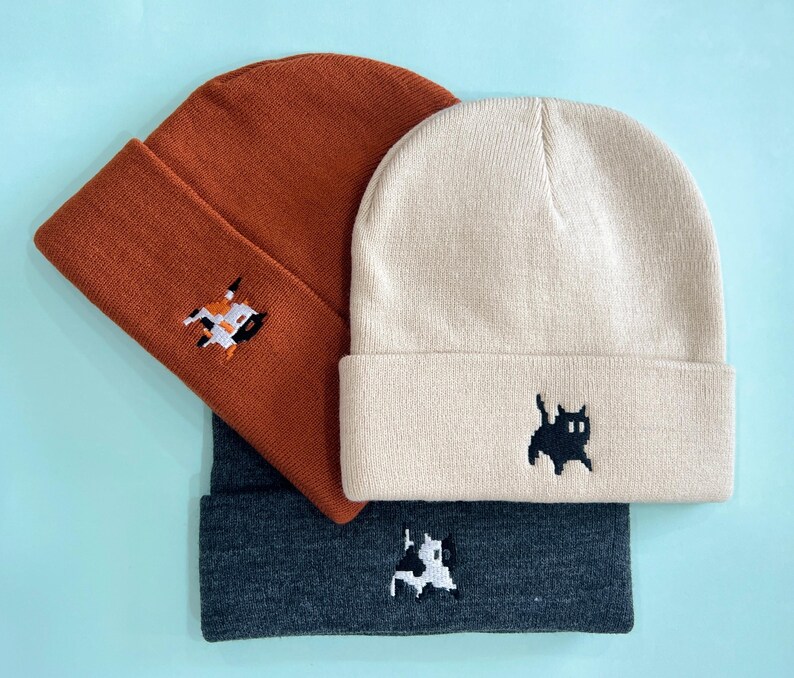 Puede incluir: Tres gorros de punto en &oacute;xido, crema y gris carb&oacute;n. Cada gorro presenta un peque&ntilde;o dise&ntilde;o bordado: un gato, una vaca y una figura geom&eacute;trica. Los gorros est&aacute;n doblados en el borde.