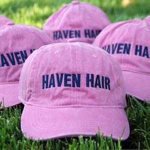 May include: Several pink baseball caps with the text "HAVEN HAIR" embroidered in navy blue. The caps are arranged on a bed of green grass, suggesting an outdoor setting. The caps have a classic six-panel design.