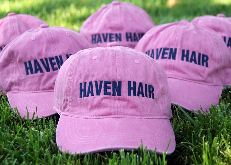 May include: Several pink baseball caps with the text "HAVEN HAIR" embroidered in navy blue. The caps are arranged on a bed of green grass, suggesting an outdoor setting. The caps have a classic six-panel design.