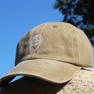 May include: A khaki green baseball cap with a white embroidered pin that says "You Are Here".