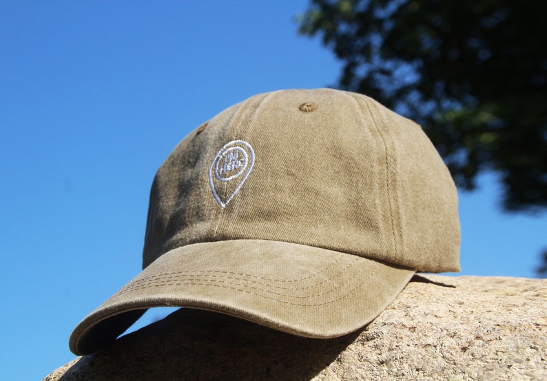 May include: A khaki green baseball cap with a white embroidered pin that says "You Are Here".