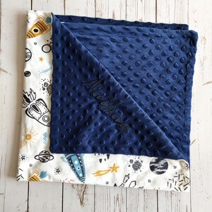 May include: A navy blue minky baby blanket with a white cotton side featuring a rocket ship and space themed print. The blanket is personalized with the name "Matthew" embroidered in black thread.