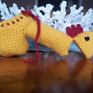 May include: A crocheted yellow chicken toy with red accents. The chicken is lying on a wooden surface.