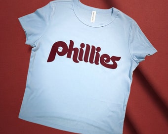 Op de afbeelding: Een lichtblauw t-shirt met een donkerrode "Phillies" logo in een retro-lettertype.