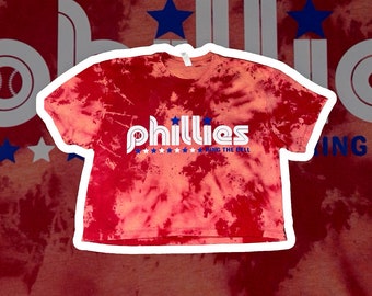 May include: Red tie-dye cropped t-shirt with a white "Phillies" logo and stars. The text below the logo reads "Ring the Bell".