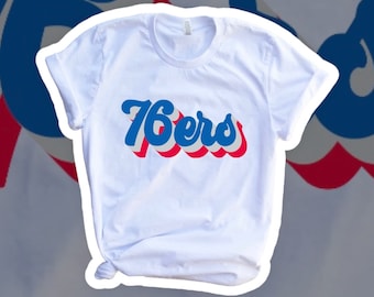 May include: White t-shirt with the text "76ers" in blue, red, and white, with a retro font. The shirt has a crew neck and short sleeves. The background has a blurred image of the 76ers logo.