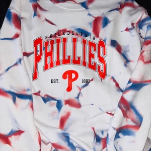 May include: White sweatshirt with a red, white, and blue tie-dye pattern. The sweatshirt has the words "Philadelphia Phillies" in red and black lettering with a red "P" logo and the text "Est. 1883" underneath.