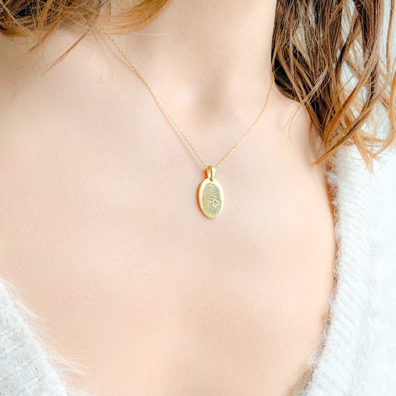 May include: A gold chain necklace with a pendant featuring a detailed fingerprint design. The pendant is oval shaped and hangs from a delicate chain.
