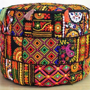Puede incluir: Un puf de suelo redondo que presenta un mosaico de paneles de tela negra bordados con vibrantes patrones geométricos multicolores, motivos florales y pequeños espejos circulares. El borde superior está decorado con varias borlas de tela.