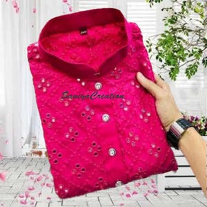Pu&ograve; includere: Una camicia rosa acceso con colletto alla coreana. La camicia presenta un motivo ricamato dettagliato con piccoli specchietti. Il testo "Saniya Creation" &egrave; visibile sulla camicia.