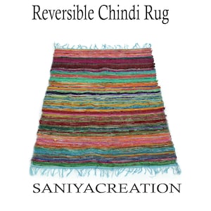 May include: Reversible Chindi rug with a vibrant, striped pattern in shades of red, green, orange, and blue. The rug has a fringed edge and the text "SANIYACREATION" at the bottom. The text "Reversible Chindi Rug" is at the top.