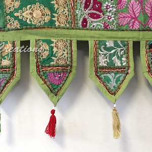 Op de afbeelding: Geborduurde textiele wandophanging in groen en goud, met een herhalend patroon van bloemen en geometrische ontwerpen. De ophanging heeft een lichtgroene rand en decoratieve kwastjes in verschillende kleuren. De tekst "Saniya Creations" is zichtbaar.