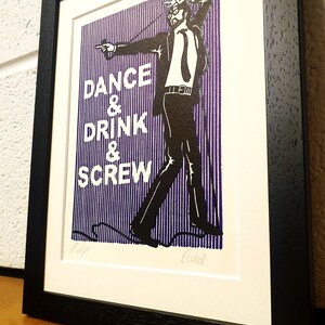 May include: Framed art print with a black frame. The print features a black and white illustration of a man in a suit with the words "DANCE & DRINK & SCREW" in white text on a purple striped background.