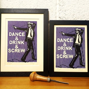 May include: Two framed art prints with black frames. The prints feature a black and white illustration of a man with the words "DANCE & DRINK & SCREW" in white text on a purple striped background. A wooden carving tool rests on the wooden surface.