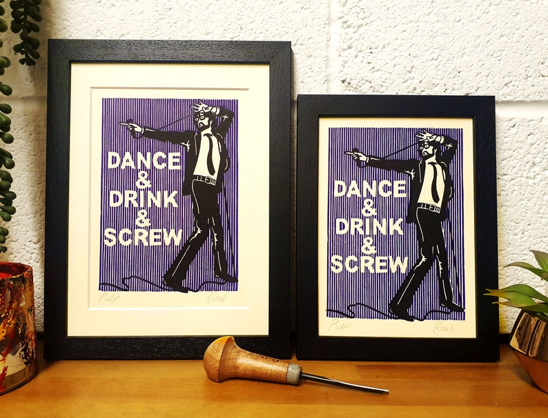 May include: Two framed art prints with black frames. The prints feature a black and white illustration of a man with the words "DANCE & DRINK & SCREW" in white text on a purple striped background. A wooden carving tool rests on the wooden surface.