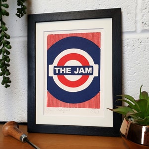 May include: A framed print of a red, white, and blue graphic design with the text "THE JAM" in a white rectangle. The graphic design is reminiscent of a London Underground sign. The print is signed "Paul"