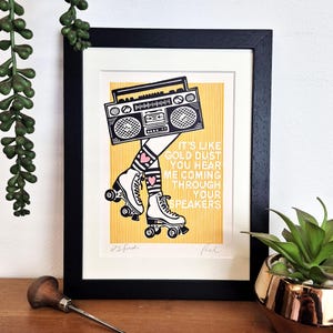 May include: A framed art print featuring a black and white illustration of a boombox head on legs wearing striped socks with pink hearts and white roller skates. The background is yellow with vertical lines and the text IT'S LIKE GOLD DUST YOU HEAR ME COMING THROUGH YOUR SPEAKERS.