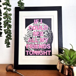 May include: A framed print featuring a black and white disco ball graphic with the pink text IF I COULD SEE ALL MY FRIENDS TONIGHT. The print is housed in a black frame with a white mat, displaying the artist signature and band name below.
