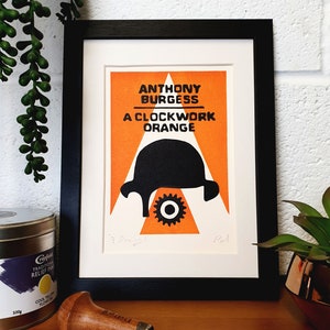 Könnte beinhalten: Ein gerahmter Druck eines Buchcovers für "A Clockwork Orange" von Anthony Burgess. Das Cover zeigt eine schwarze Silhouette eines menschlichen Kopfes mit einem Zahnrad in der Mitte, vor einem leuchtend orangen Hintergrund mit weißen Akzenten.