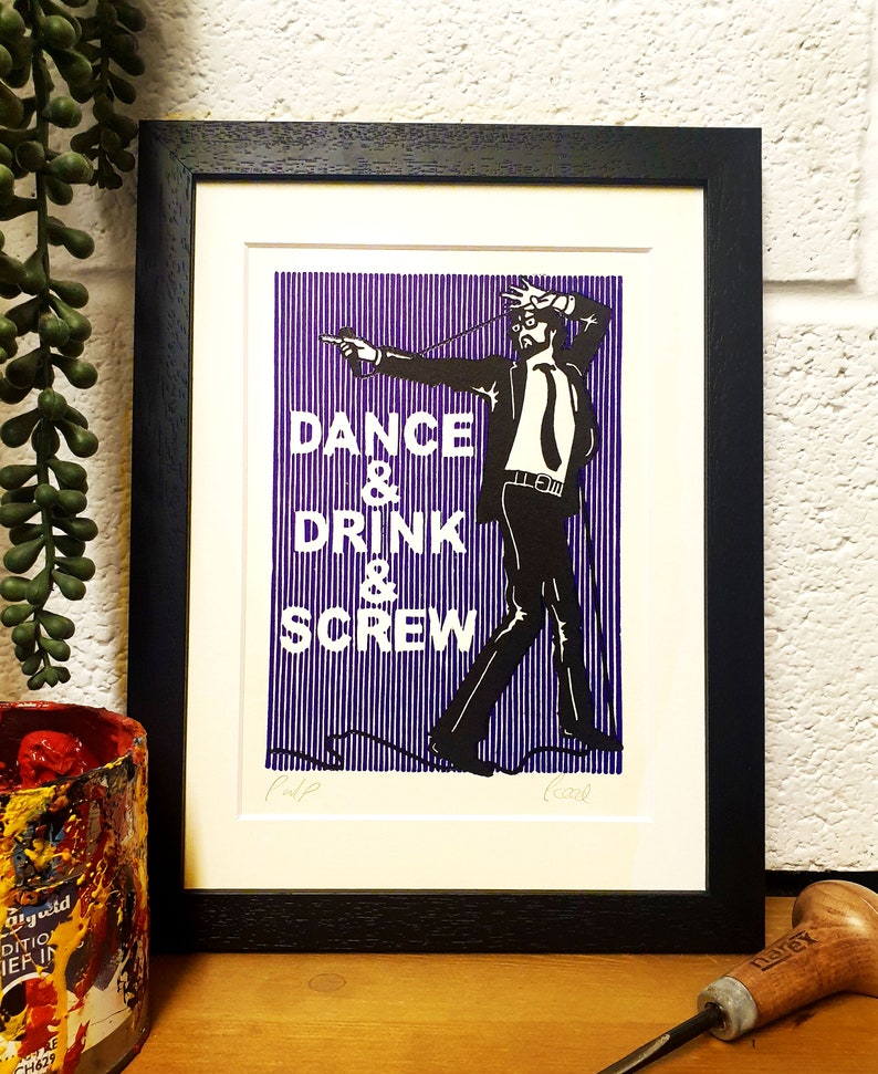 May include: Framed art print with a black frame. The print features a black and white illustration of a man in a suit with the words "DANCE & DRINK & SCREW" in white. The background is vertical blue and white stripes.