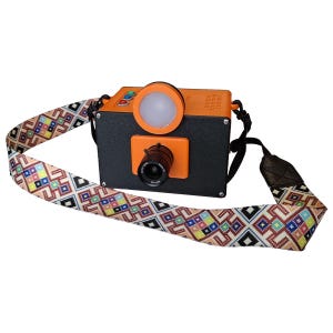 May include: A rectangular camera with a black front panel, an orange top, and a circular lens. It features a round white light, three small colored buttons on top, and a long, wide camera strap decorated with a colorful geometric pattern.