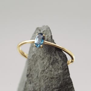 Könnte beinhalten: Ein zarter Goldring mit einem kleinen, ovalförmigen blauen Edelstein. Der Ring hat ein dünnes, poliertes Band, und der Edelstein ist in einer einfachen Vier-Krappen-Fassung gefasst. Der Ring wird auf einer grauen, strukturierten Oberfläche präsentiert.