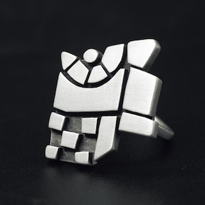 Könnte beinhalten: Ein silberfarbener Ring mit geometrischem Design. Der Ring weist eine Reihe von rechteckigen und quadratischen Formen auf, die in einem geschichteten Muster angeordnet sind. Der Ring hat eine moderne, abstrakte Ästhetik mit glatter, polierter Oberfläche.
