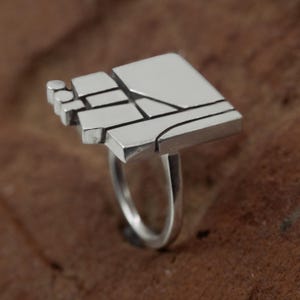 Könnte beinhalten: Ein silberner Ring mit geometrischem Design. Der Ring hat eine rechteckige Oberseite mit einer Reihe von geschichteten Formen und Linien. Das Band ist ein einfaches, glattes Silber. Der Ring ist vor einem braunen Hintergrund platziert.