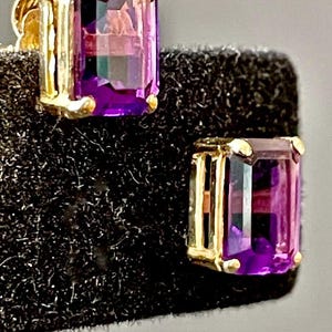 May include: A pair of gold earrings with purple amethyst gemstones. The gemstones are emerald cut and set in a bezel setting.