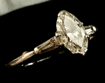 Puede incluir: Un anillo de plata con un gran diamante ovalado transparente engastado en garras. El diamante está rodeado de diamantes más pequeños.