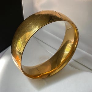 May include: A wide, polished gold-tone bangle bracelet with a smooth, reflective surface. The circular accessory features a seamless design with a visible hinge closure, showcasing a bright, metallic finish that catches the light.