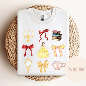 Puede incluir: Sudadera blanca con ilustraciones en acuarela de un vestido amarillo, lazos rojos, libros, un espejo, una taza de té y un candelabro. El diseño también incluye un lazo amarillo. La sudadera se muestra sobre una cesta tejida.