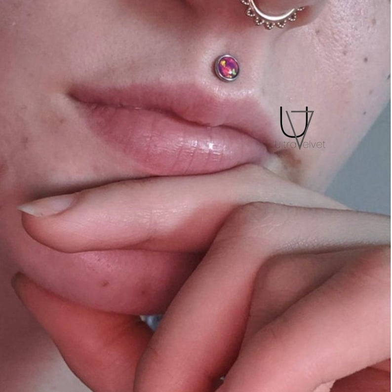 Puede incluir: Primer plano de la parte inferior de la cara de una persona con un peque&ntilde;o piercing de &oacute;palo en el labio. El piercing es un pendiente de plata con una piedra de &oacute;palo redonda de color rosa, naranja y amarillo.