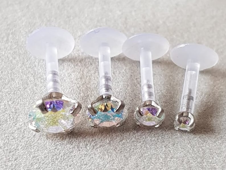 Op de afbeelding: Vier transparante lichaamssieraden van plastic met regenboogkleurige edelstenen. De stenen zijn rond en hebben een regenboogeffect.