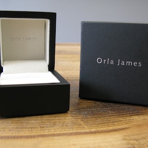 May include: A black square box with a white interior and the text "Orla James" on the lid. The box is open and the interior is visible.