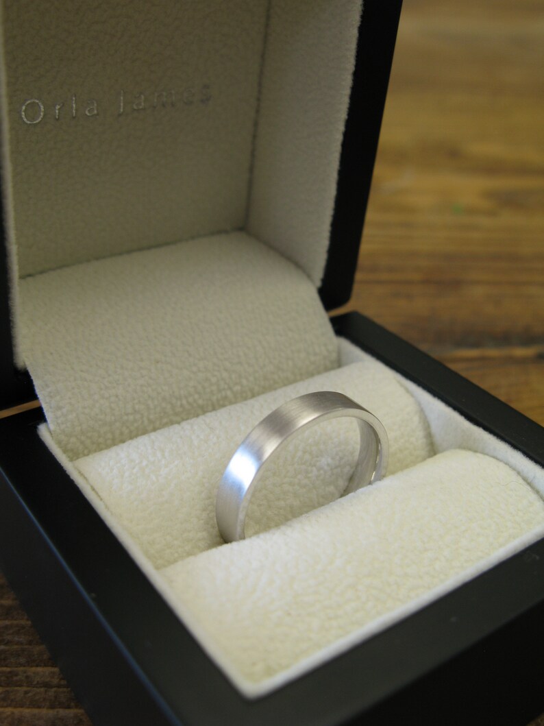 May include: A silver wedding band in a black ring box with a white interior. The box has the text "Orla James" on the lid.