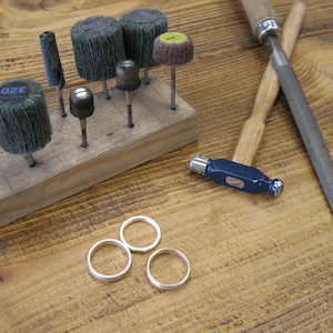 May include: A wooden workbench with a variety of tools, including sanding discs, a saw, a file, and a hammer. There are also three silver rings on the workbench.