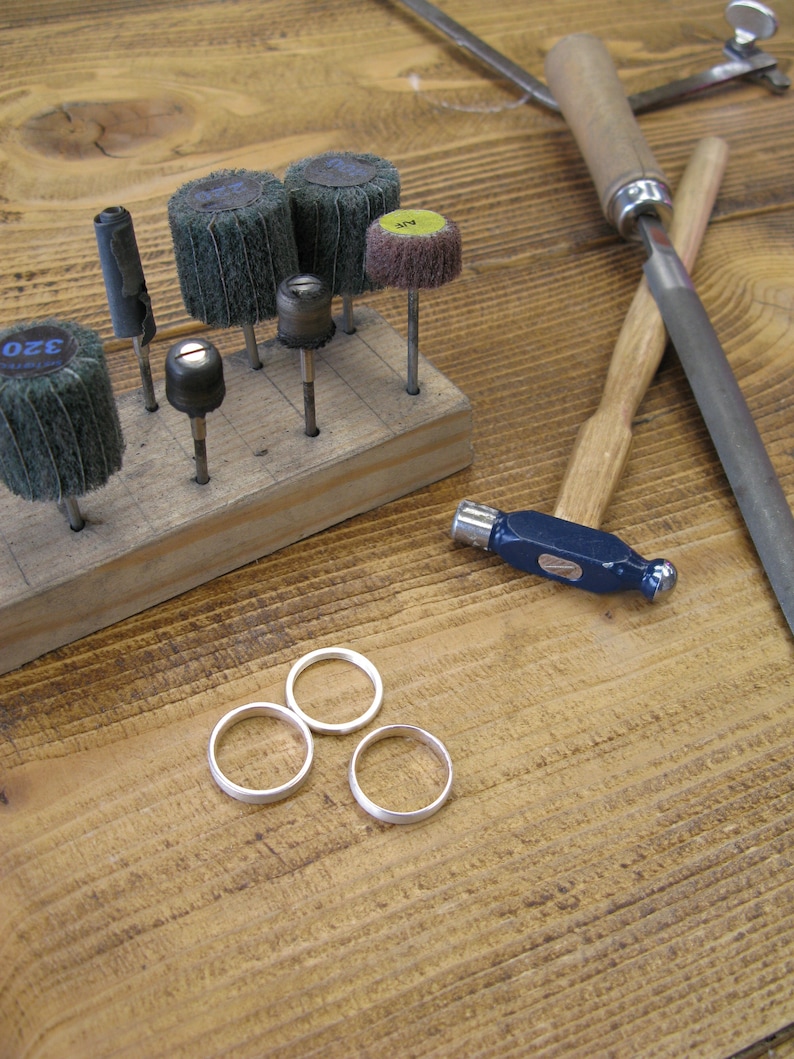May include: A wooden workbench with a variety of tools, including sanding discs, a saw, a file, and a hammer. There are also three silver rings on the workbench.