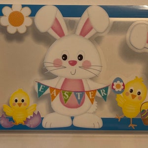 May include: Easter-themed decorative frame with white bunnies, colorful eggs, and yellow chicks. The word "EASTER" is displayed on a banner. The frame has a blue border and is perfect for spring holiday decorations.