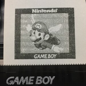 May include: A black and white image of a Nintendo Game Boy cartridge with a pixelated image of Mario on the front. The text "Nintendo" and "GAME BOY" are printed on the cartridge.