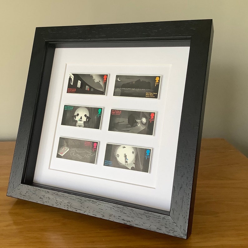 May include: A black framed display of eight vintage postage stamps featuring illustrations from the novel "The Murder of Roger Ackroyd" by Agatha Christie. The stamps are arranged in a grid pattern and feature a variety of scenes from the book, including a skull, a house, and a figure in a boat.