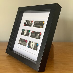 May include: A black framed picture with a white mat and six black and white stamps inside. The stamps feature images of a skull, a person, a building, and a landscape. The stamps are all from the same series and have the same design.