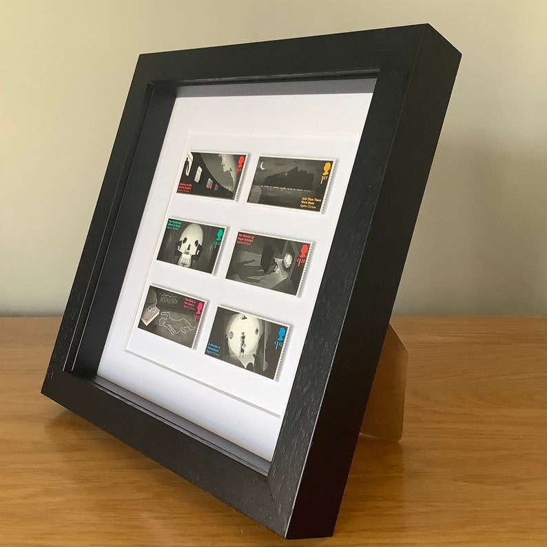 May include: A black framed picture with a white mat and six black and white stamps inside. The stamps feature images of a skull, a person, a building, and a landscape. The stamps are all from the same series and have the same design.