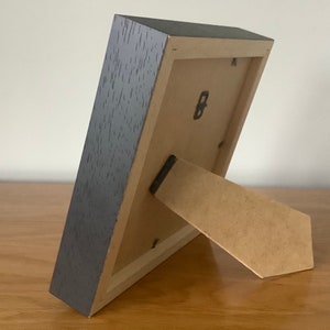 May include: A dark gray wood picture frame with a cardboard easel stand on the back. The frame has a small metal hook on the back.