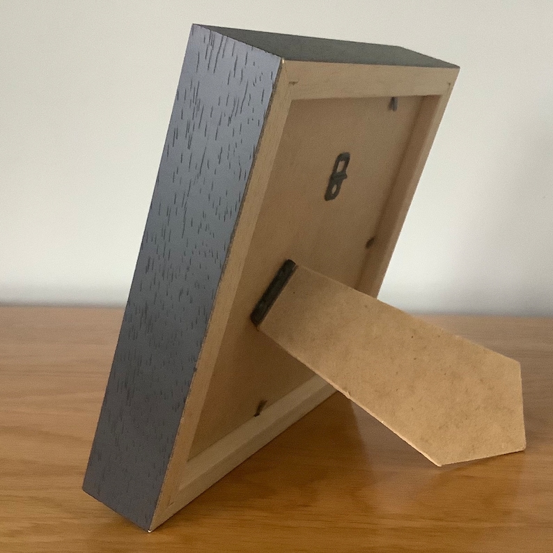 May include: A dark gray wood picture frame with a cardboard easel stand on the back. The frame has a small metal hook on the back.
