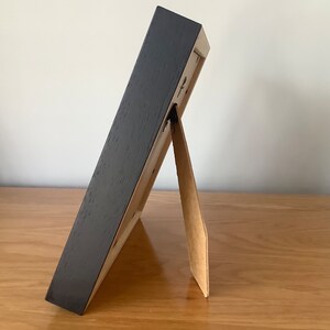 May include: A black picture frame with a brown cardboard easel stand. The frame is standing on a wooden surface.