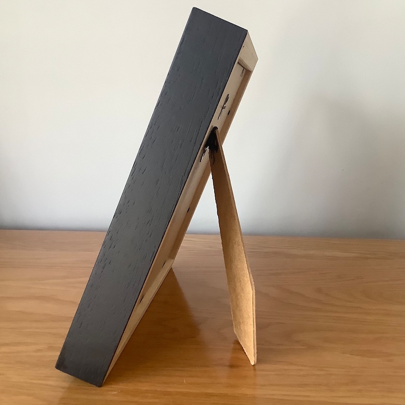 May include: A black picture frame with a brown cardboard easel stand. The frame is standing on a wooden surface.