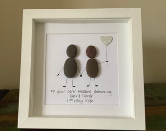 May include: A framed pebble art piece depicting two figures holding hands with a silver heart balloon. The text reads "On your Silver Wedding Anniversary Kim & Trevor 11th May 1996".