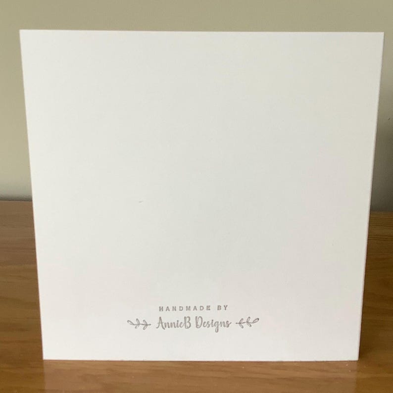 Puede incluir: Una tarjeta blanca con el texto "Handmade by Annie B Designs" en letras grises.