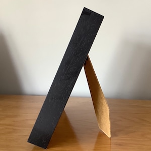 May include: A black picture frame with a cardboard easel stand. The frame is made of wood and has a smooth finish.